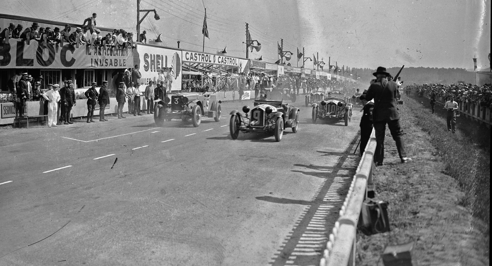 Départ des 24 Heures du Mans 1932, photo d’archive en noir et blanc : les voitures quittent la ligne devant les stands.